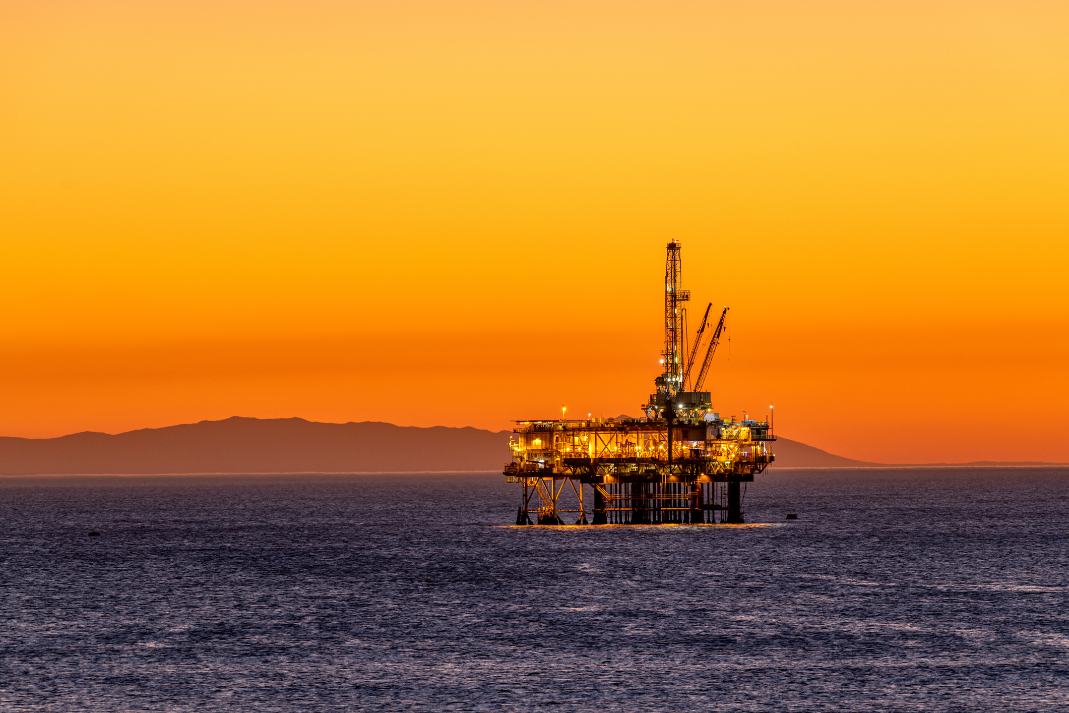 Offshore oil platform off the coast of California against a moody, orange sky as the sun sets behind the rig.