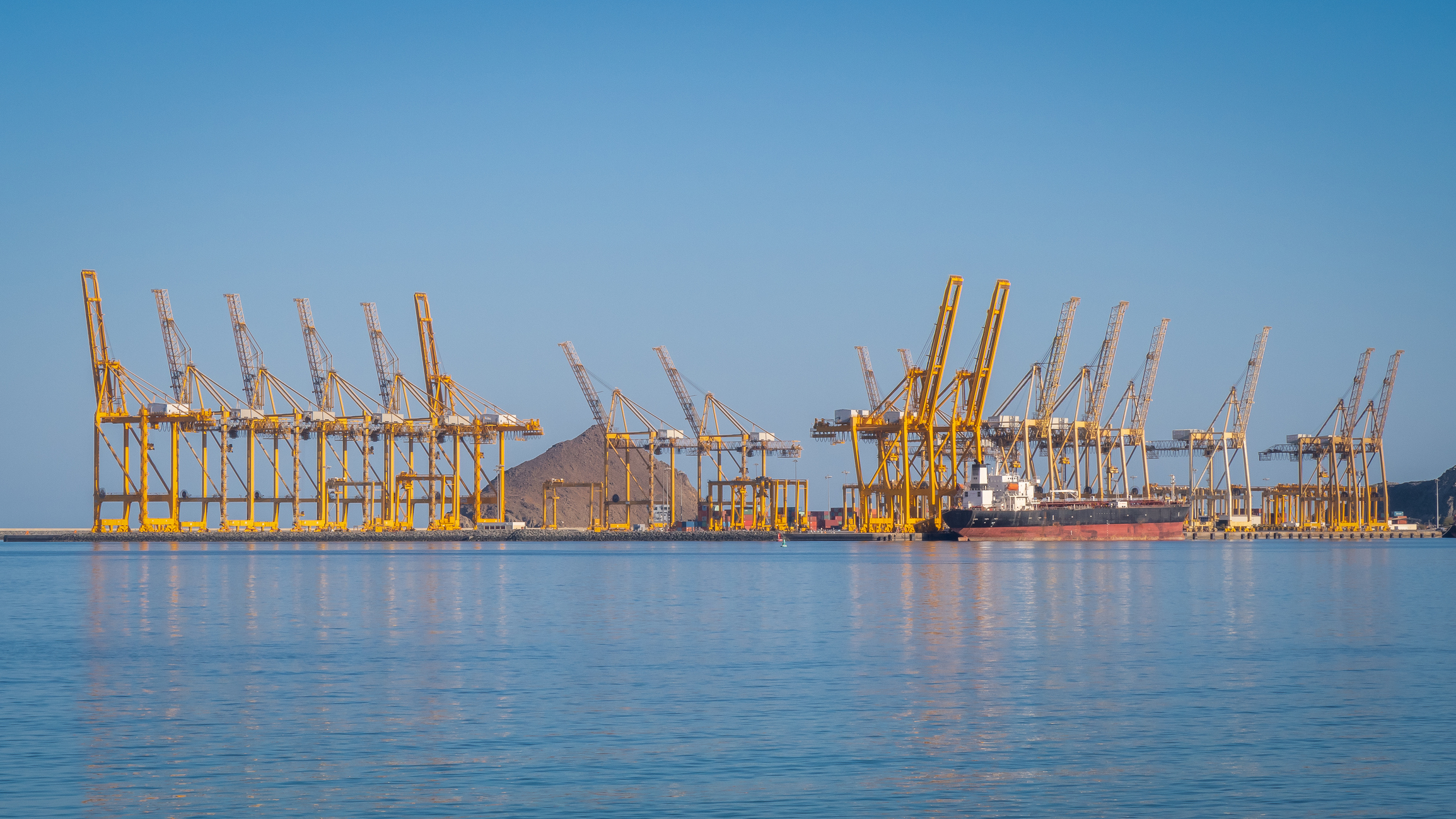 Port cranes loading container ship in the Port of Fujairah, United Arab Emirates. Rows of yellow port cranes against blue sea and sky.